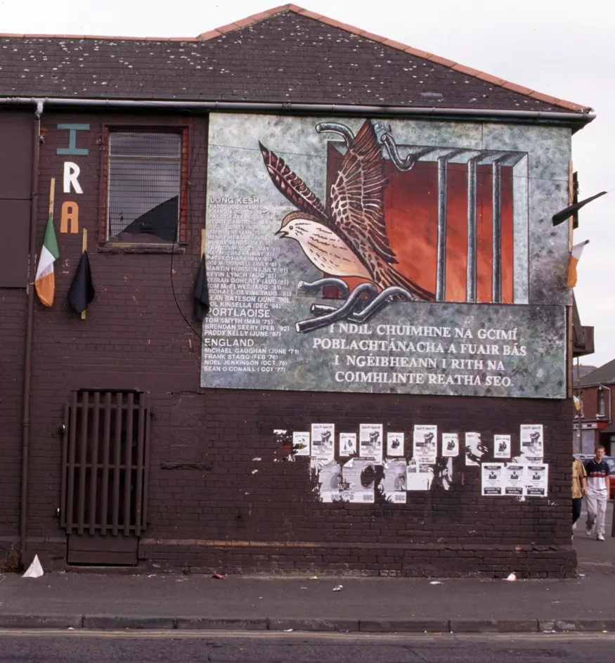 Cartoon mural in Catholic Falls Road area of Belfast depicting a caged bird escaping from behind bars.
Inscription reads (In Irish), ""IRA / I NDIL CHUIMHNE NA GCIMI POBLACHTANACHA A FUAIR BAS I NGEIBHEANN I RITH NA COIMHLINTE REATHA SEO.""
