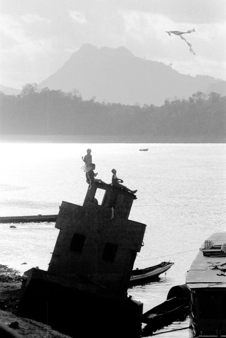 Children fly a kite from the top of an old wrecked boat along the shore of the Mekong River.